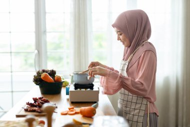 Modern mutfakta tesettürlü, mutlu yemek yapan, tencereye tahta kaşıkla yemek yiyen, taze sebzeler ve meyveler yerleştiren Müslüman bir kadın. Kepçelemek ve yemeği tatmak keyifli..