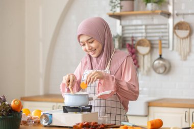 Modern mutfakta tesettürlü, mutlu yemek yapan, tencereye tahta kaşıkla yemek yiyen, taze sebzeler ve meyveler yerleştiren Müslüman bir kadın. Kepçelemek ve yemeği tatmak keyifli..