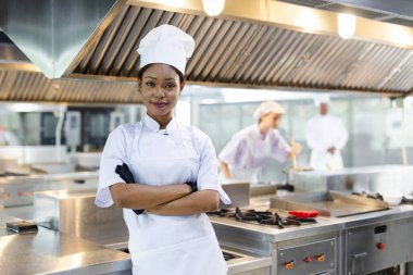 Portre şefi aşçılık uzmanı kadınlar, donanımlı mutfakta kollarını kavuşturup, güven ve profesyonellik gösterirken, iş arkadaşlarının arkasında yemek pişirdiği belli oluyor..