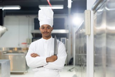 Usta şefin portresi mutfak arka planında duruyor, kolları çaprazlanmış beyaz üniforma ve şef şapkası takıyor..
