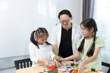 Gülümseyen öğretmen iki kıza renkli STEM robotik projeleri inşa etmede, yaratıcılığı teşvik etmede, problem çözmede ve ekip çalışmasında yardımcı oluyor..