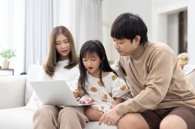 Evde dizüstü bilgisayar kullanan Asyalı bir aile, koltukta ebeveynler arasında oturan bir kız, ekran başında öğrenme ve eğlenme, modern ebeveynlik, kaynaşma ve dijital aile yaşam tarzını gösteren.