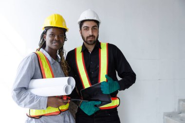 Güvenlik teçhizatı kullanan Afrikalı kadın ve Ortadoğulu erkek inşaat mühendisleri kameraya bakıyor, ellerinde planlar ve tablet, takım çalışması, çeşitlilik ve iş sahası işbirliğini temsil ediyorlar..