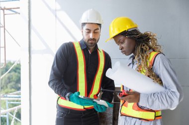 Güvenlik miğferleri ve yansıtıcı yeleklerde inşaat mühendisleri tablet ve planlar kullanarak bina planlarını gözden geçirir, inşaat yönetimindeki takım çalışması, planlama ve dijital araçların altını çizer.