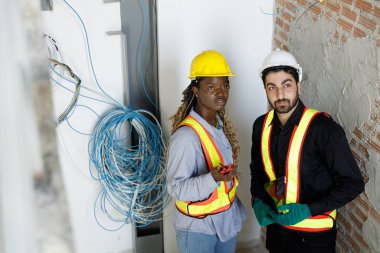 İki inşaat mühendisi şebeke kablosu kurulumunu inceliyor. Güvenlik teçhizatı ile, korunmasız kablolar ve altyapının yanında dururken bir tablet ve ölçüm aracı tutuyorlar..