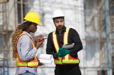 İki inşaat mühendisi, telsizle konuşurken tabletteki proje verilerini gözden geçiriyorlar. Açık hava inşaat ortamında güvenlik, planlama ve takım çalışması kavramı.