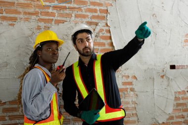 İki inşaat işçisi inşaat planlarını tartışıyorlar. Güvenlik yelekleri ve miğferler giyerek, dijital tabletle ilgili belge ve verileri gözden geçiriyorlar.