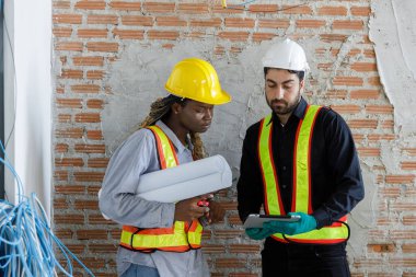 İki inşaat işçisi inşaat planlarını tartışıyorlar. Güvenlik yelekleri ve miğferler giyerek, dijital tabletle ilgili belge ve verileri gözden geçiriyorlar.