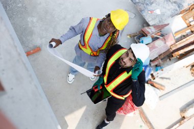 Güvenlik kaskı takan iki inşaat mühendisi ve inşaat alanındaki planları tartışan yansıtıcı yeleklerle takım çalışmasını ve planlamasını temsil ediyorlar. Yukarıdan görüntüle.