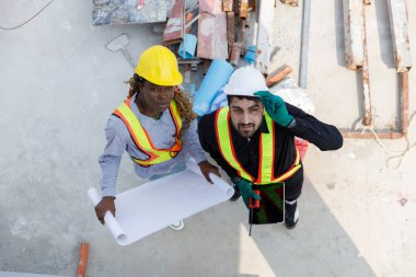 Güvenlik kaskı takan iki inşaat mühendisi ve inşaat alanındaki planları tartışan yansıtıcı yeleklerle takım çalışmasını ve planlamasını temsil ediyorlar. Yukarıdan görüntüle.