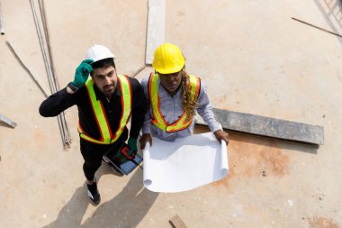 Güvenlik kaskı takan iki inşaat mühendisi ve inşaat alanındaki planları tartışan yansıtıcı yeleklerle takım çalışmasını ve planlamasını temsil ediyorlar. Yukarıdan görüntüle.