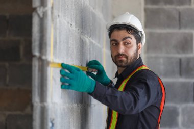 Erkek inşaat işçisi, tuğla duvarı ve mezuranın ölçüsünü alıyor. Güvenlik yeleği, sarı kask ve eldiven giymek, inşaat işlerinde hassasiyeti, işçiliği ve saha denetimini temsil eder..