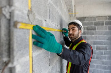 Erkek inşaat işçisi, tuğla duvarı ve mezuranın ölçüsünü alıyor. Güvenlik yeleği, sarı kask ve eldiven giymek, inşaat işlerinde hassasiyeti, işçiliği ve saha denetimini temsil eder..