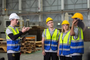 Fabrikadaki brifing sırasında denetleyiciyi dinleyen güvenlik yelekleri ve miğferler içinde çeşitli endüstriyel işçiler, takım çalışması, eğitim, disiplin ve iş güvenliği iletişimini gösteriyor..