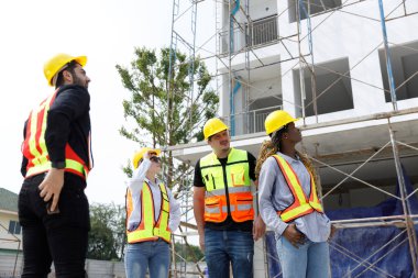 İnşaat alanındaki ilerlemeyi denetleyen, takım çalışması, mühendislik, güvenlik ve proje geliştirme planlamasını temsil eden güvenlik kaskı ve yansıtıcı yelek giyen çeşitli inşaat işçileri grubu.