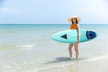 Tropik plajda elinde sörf tahtası, gülümseyen genç bir kadın. Yaz tatili kavramı, sörf yaşam tarzı, açık hava aktivitesi, özgürlük ve plaj macerasından zevk alan kadın gezgin..