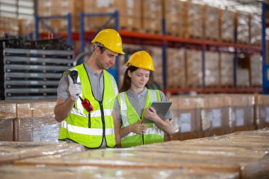 Depo çalışanları güvenlik yelekleri ve miğferleriyle envanter kontrolü yapıyor ve dijital tabletleri stok yönetimi için kullanıyor. Modern lojistik ve sanayi operasyonlarında takım çalışması.