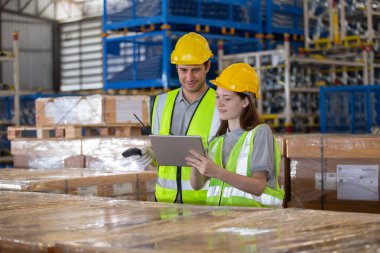 Depo çalışanları güvenlik yelekleri ve miğferleriyle envanter kontrolü yapıyor ve dijital tabletleri stok yönetimi için kullanıyor. Modern lojistik ve sanayi operasyonlarında takım çalışması.