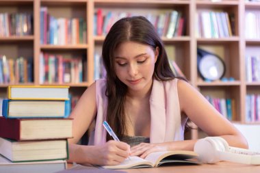 Bir kitap yığınıyla kütüphanede okuyan genç bir kadın, sessiz ve üretken bir ortamda eğitim, araştırma veya akademik hazırlık notları yazmaya odaklanmış..