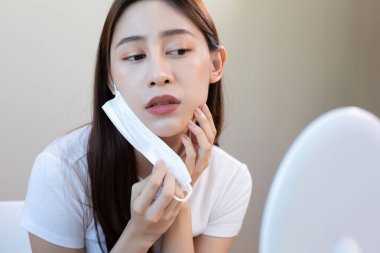 Asyalı kadının yüz maskesi çıkarması ve aynada sivilce kontrolü yapması endişe verici, maske sorununu temsil ediyor, cilt sorunu ve uzun süreli maske kullanımının neden olduğu yüz tahrişi..