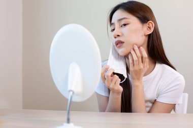 Asyalı kadının yüz maskesi çıkarması ve aynada sivilce kontrolü yapması endişe verici, maske sorununu temsil ediyor, cilt sorunu ve uzun süreli maske kullanımının neden olduğu yüz tahrişi..