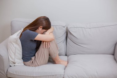 Asyalı bir kadın koltukta tek başına oturuyor, başı kollarına eğik, üzüntüsünü ifade ediyor, duygusal stres, zihinsel sağlık sorunu, ya da yalnızlık ve depresyon hissi..