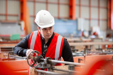 Beyaz güvenlik kaskı ve kırmızı yansıtıcı yelek giyen endüstri mühendisi, fabrikada ağır makineler çalıştırıyor. Odaklanma, hassasiyet ve ağır fabrikada çalışmanın altını çiziyor..