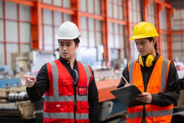 Güvenlik yelekleri ve miğferli iki sanayi işçisi fabrikadaki panodaki kontrol listesini incelerken operasyonları tartışıyorlar. Takım iletişimi, planlama ve işyeri güvenliği kavramı.