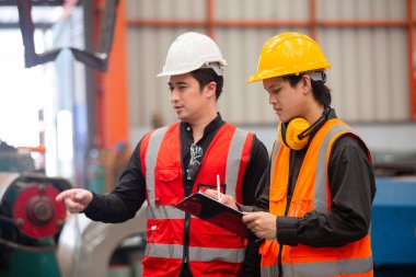 Güvenlik yelekleri ve miğferli iki sanayi işçisi fabrikadaki panodaki kontrol listesini incelerken operasyonları tartışıyorlar. Takım iletişimi, planlama ve işyeri güvenliği kavramı.