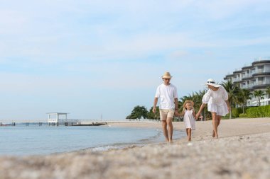 Tropikal sahilde çıplak ayakla yürüyüp el ele tutuşan ve gülümseyen mutlu bir aile. Baba, anne ve kız birlikte yaz tatilinin tadını çıkarıyorlar. Aile bağı, yaşam tarzı, aşk ve özgürlük kavramı.