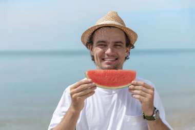 Hasır şapkalı ve beyaz gömlekli gülümseyen adam tropik plajda dilimlenmiş karpuz tutuyor. Yaz tatili kavramı, sağlıklı beslenme, meyve ferahlığı, tropikal yaşam tarzı ve mutluluk..