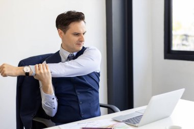 Resmi takım elbiseli bir işadamı dizüstü bilgisayarla masada otururken kollarını uzatıyor, ofisin rahatlığını, stres atılımını ya da modern kurumsal çalışma alanındaki sağlıklı çalışma alışkanlıklarını gösteriyor..