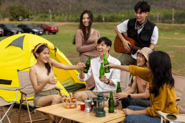 Bir grup Asyalı genç arkadaş göl kenarında kamp yapıyor, içkilerini paylaşıyor, gitar çalıyor ve eğlenceli ve rahatlatıcı bir atmosferde birlikte yemek pişiriyorlar..