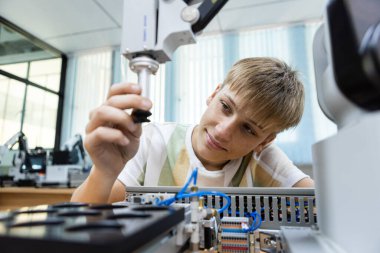 Okulun teknoloji laboratuvarında robot kolu test eden gülümseyen bir öğrenci. STEM eğitimi, robotik öğrenme, mühendislik, otomasyon sistemleri ve teknolojide gençlik yeniliği kavramı.