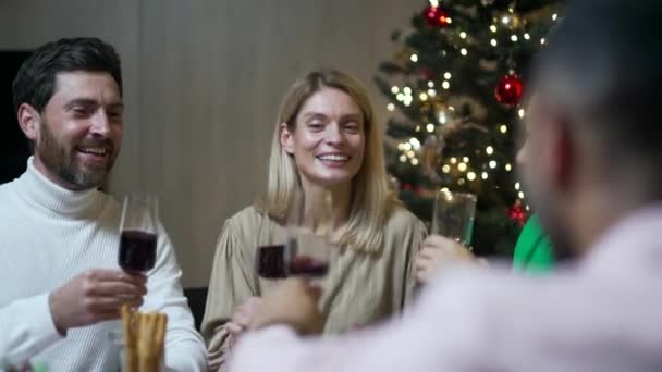 友人は一緒にテーブルに座ってクリスマスの前夜または新年の休日のお祝いを祝います 多民族のお祝いのクリスマスディナーのホームグループでの饗宴 幸せな陽気な人々クリンクワイングラスは笑う — ストック動画