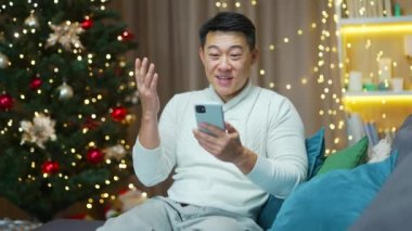 Heyecanlı Asyalı genç adam mutlu mutlu bir gülümseme zaferi kutla akıllı telefon xmas kış evde oturup Noel arifesinde cep telefonuna bakmak sürpriz oldu cep telefonundan harika haberler aldım