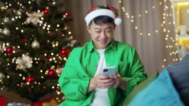 Noel Baba şapkalı neşeli, komik Asyalı adam yılbaşında evde oturup akıllı telefon kullanıyor. İyi okumalar. Komik videolar, sohbetli cep telefonları. Gülüyor.