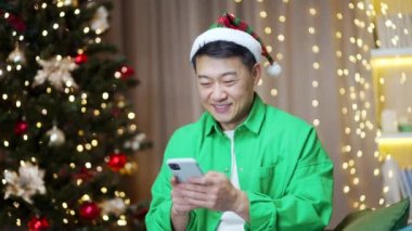 Noel Baba şapkalı neşeli, komik Asyalı adam yılbaşında evde oturup akıllı telefon kullanıyor. İyi okumalar. Komik videolar, sohbetli cep telefonları. Gülüyor.