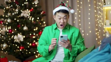Heyecanlı Asyalı genç adam mutlu mutlu bir gülümseme zaferi kutla akıllı telefon xmas kış evde oturup Noel arifesinde cep telefonuna bakmak sürpriz oldu cep telefonundan harika haberler aldım