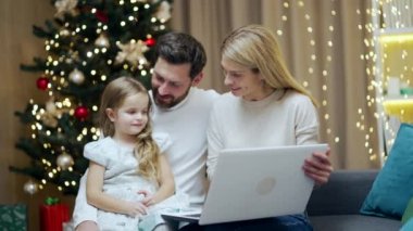Genç ebeveynler çocuklarına dizüstü bilgisayar kullanmayı internetten internet öğrenmeyi, oturma odasındaki rahat koltukta oturmayı Noel ağacının arka planında oturmayı, anne ve babalarının küçük kızları olmasını öğretiyor.