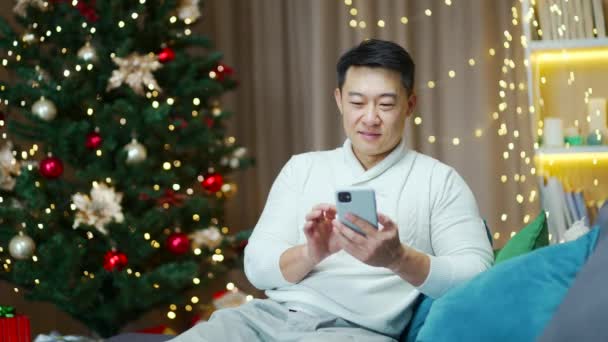 アジアの男性は クリスマスツリーに対する冬の新しい年の休日の間にソファの上に自宅に座ってスマートフォンを使用していますビジネス男性フリーランス携帯電話のブラウジングチャット居心地の良いアパートで買い物を入力 — ストック動画
