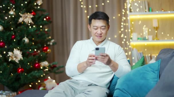 Asiatischer Mann Sitzt Den Winterferien Hause Auf Sofa Gegen Weihnachtsbaum — Stockvideo