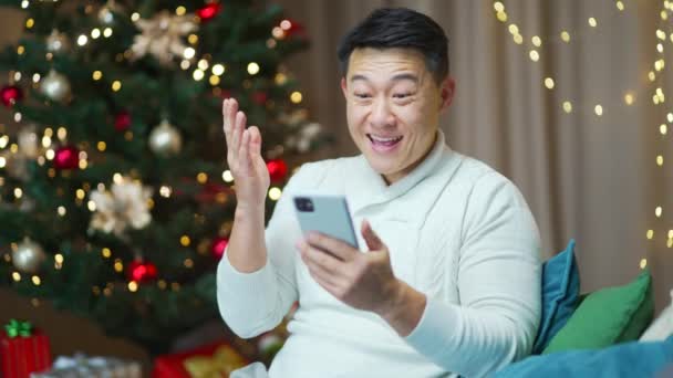 若い興奮したアジアの男幸せな肯定的な笑顔は自宅で座って勝利のスマートフォンXmas冬を祝う喜びます屋内で驚いている携帯電話を見てクリスマスイブ携帯電話で素晴らしいニュースを受け取る — ストック動画