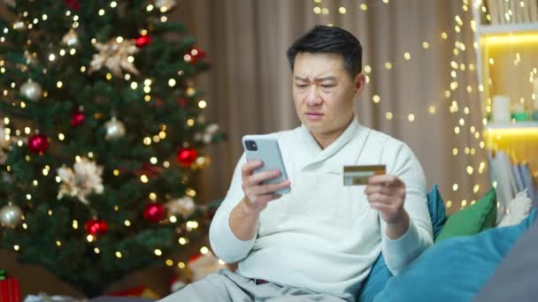 支払いの難しさ オンラインストアで購入しようとしている不満アジアの男 男はクリスマスにだまされました 自宅でスマートフォンとクレジットカードでソファに座って — ストック動画