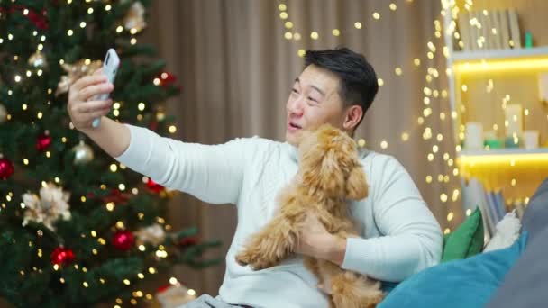 亚洲男人和宠物狗一起庆祝新年 他们用智能手机打电话问候新年 还坐在家里过着快乐的圣诞节 在客厅里的沙发上摆着圣诞装饰品 — 图库视频影像