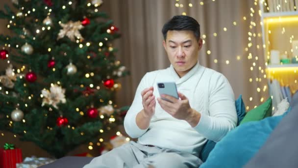 心配アジアの男は携帯電話のスマートフォンの画面を見て大晦日の日に悪いニュースメッセージ通知を読んで失望絶望男性は家庭の背景クリスマスツリーに座って — ストック動画