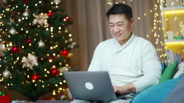 ハンサムなアジアの男はクリスマスツリーに対して冬の新年の休日の間にソファの上に自宅に座ってラップトップコンピュータを使用します — ストック動画