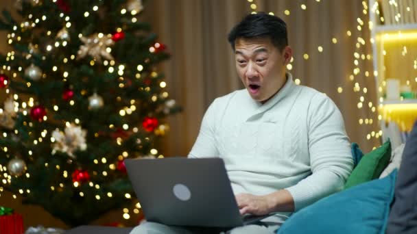 兴奋的亚洲人欣喜若狂地庆祝伟大的在线新闻 坐在沙发上看着笔记本电脑屏幕上的成功 享受商业报价 赢了圣诞节 — 图库视频影像