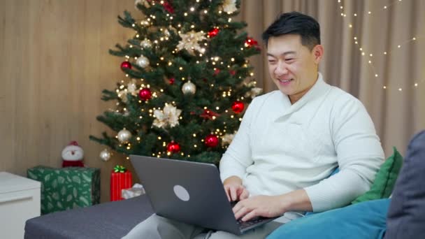 ハンサムなアジアの男はクリスマスツリーに対して冬の新年の休日の間にソファの上に自宅に座ってラップトップコンピュータを使用します — ストック動画