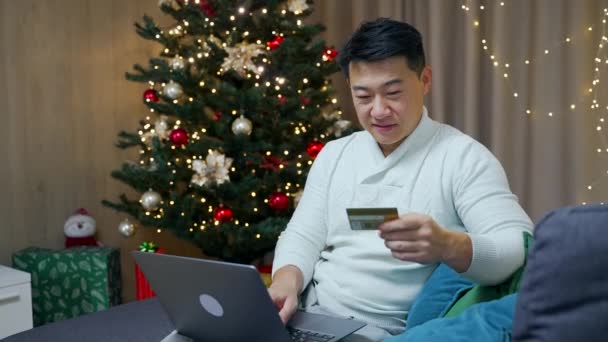 在圣诞树寒假期间 亚洲男人坐在沙发上 用笔记本电脑和信用卡在网上购物 快乐的男性支付礼物使得网上订单支付使用计算机 — 图库视频影像